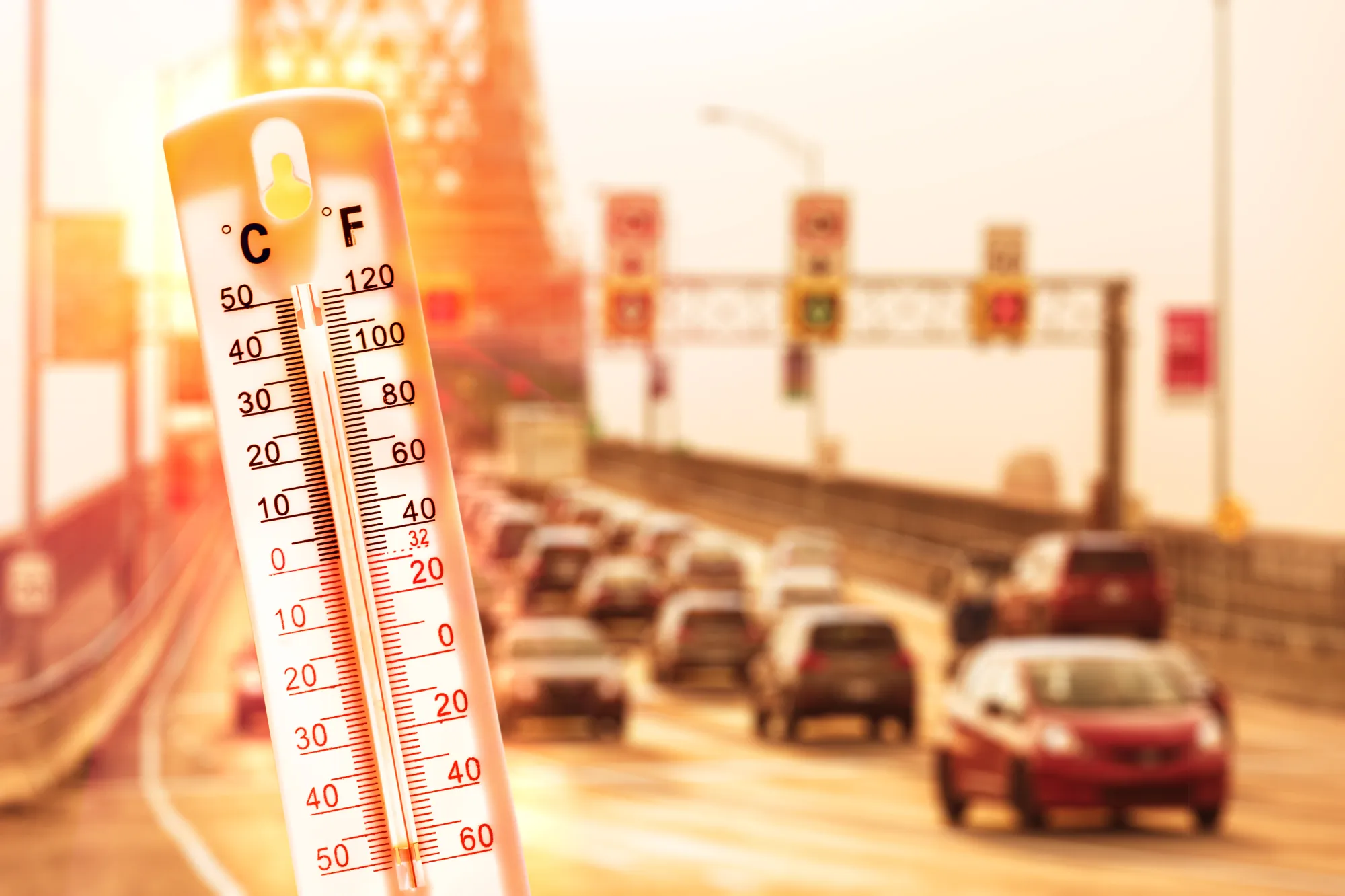 A thermometer showing a high temperature in front of a heat-hazed highway filled with cars.