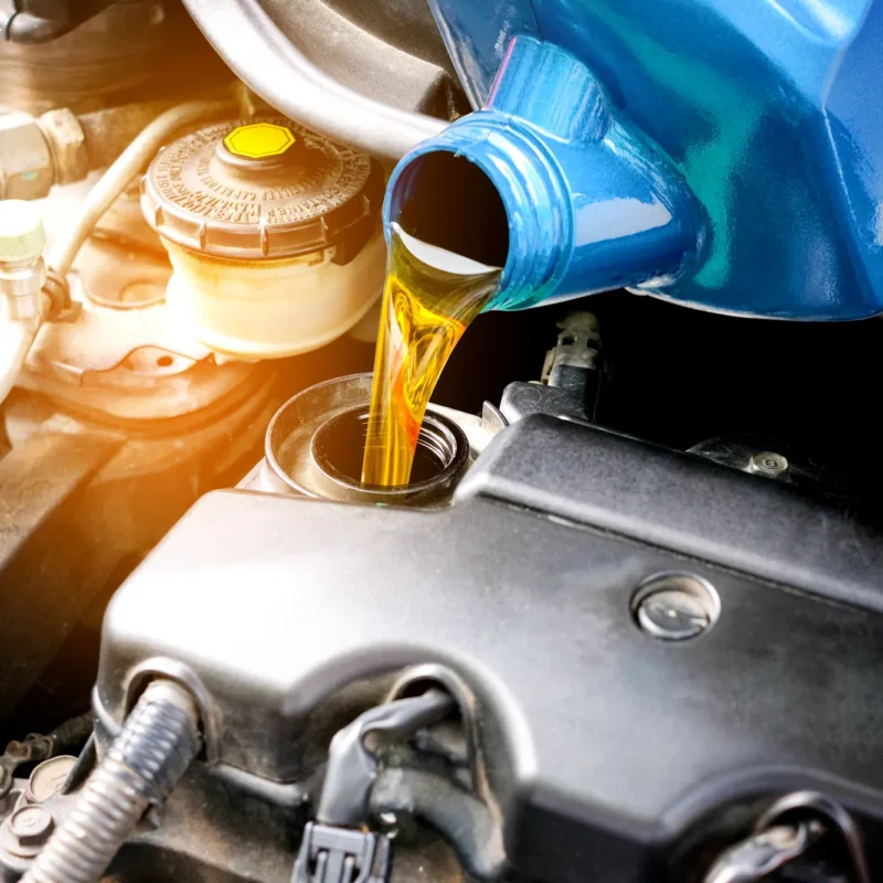 Golden motor oil pouring from a blue bottle into a car engine's oil filler opening under the hood.