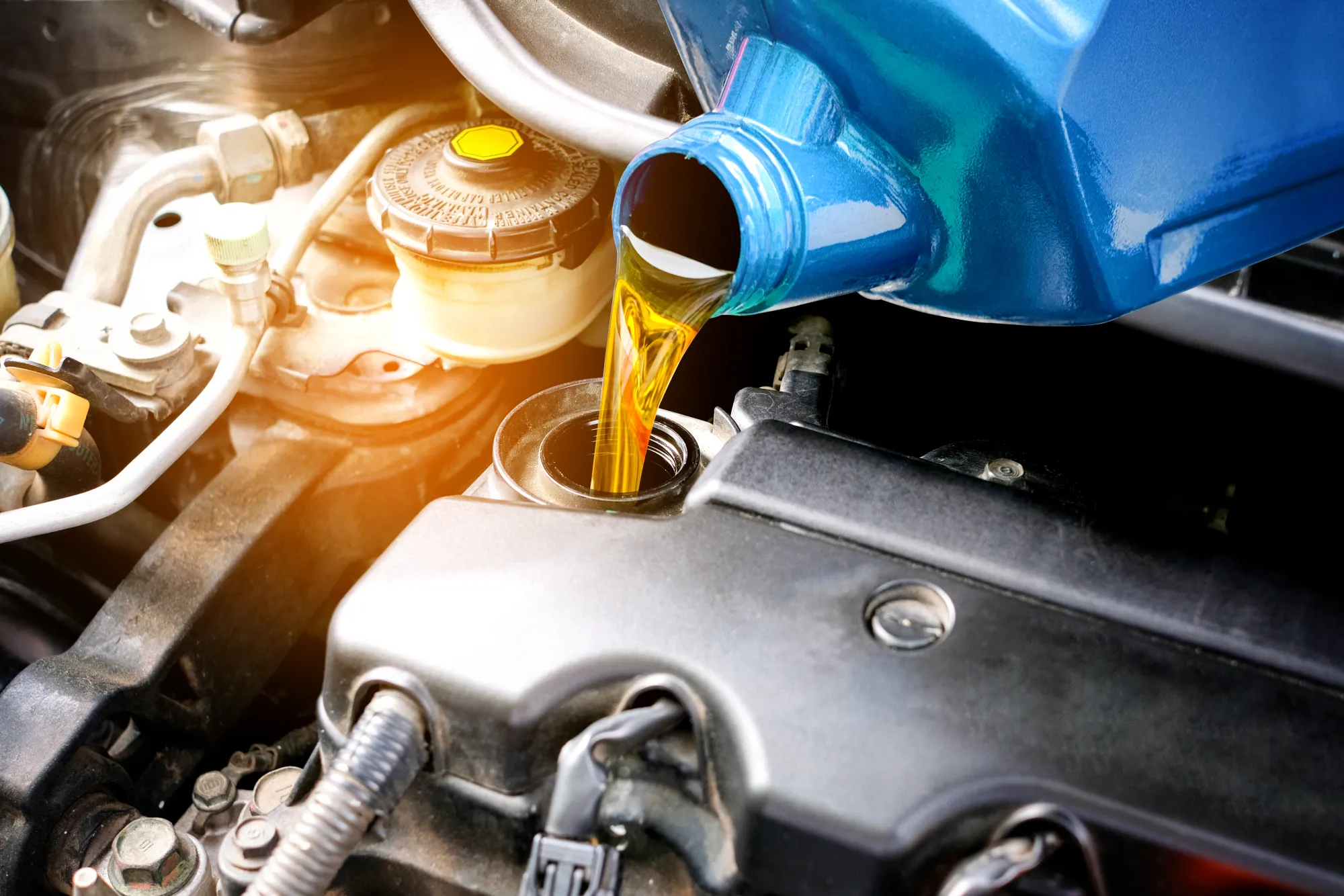 Golden motor oil pouring from a blue bottle into a car engine's oil filler opening under the hood.